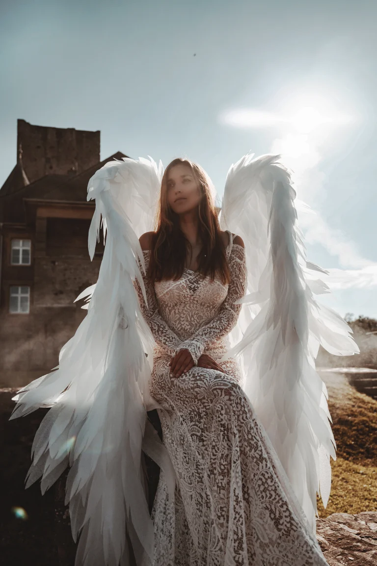 Outdoor fine art portrait of a woman in a lace gown with angel wings, photographed near a castle in soft sunlight.