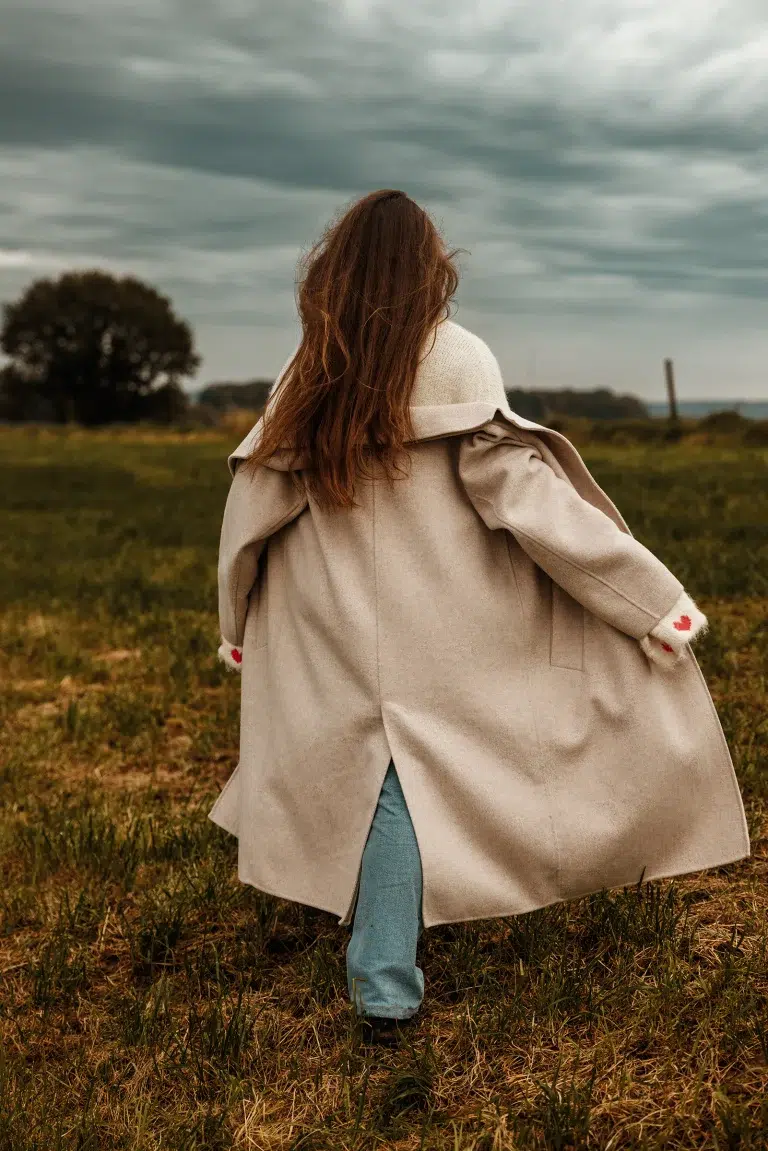 Candid outdoor photo of a woman walking through a field in a beige coat and jeans under a cloudy sky.