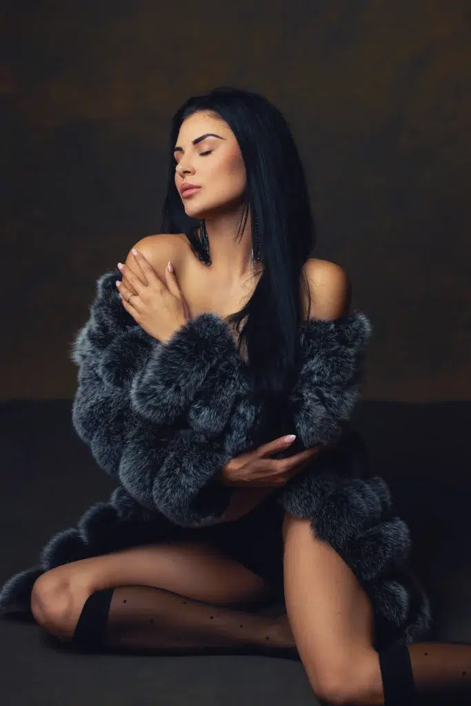 Boudoir portrait of a dark-haired woman wearing a grey fur coat, kneeling in a warm-toned studio, Massen Photography Luxembourg