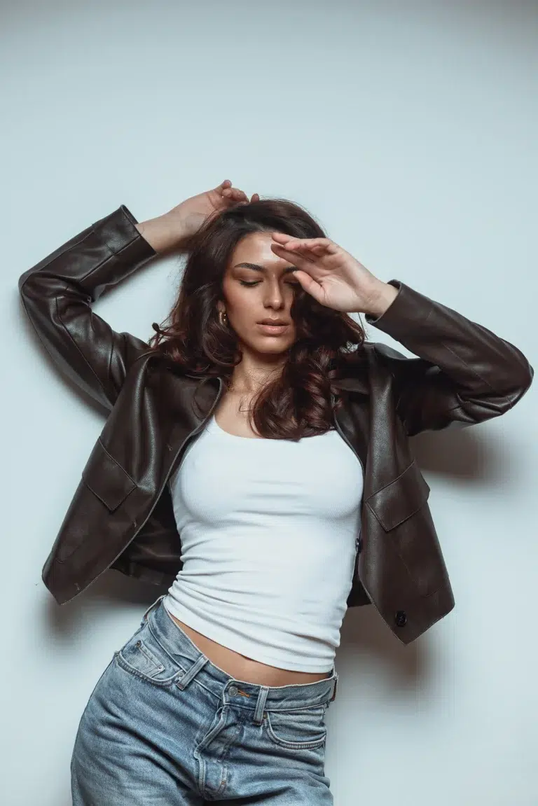 Woman in a brown leather jacket and white top leaning against a light wall, eyes closed, one hand raised to her forehead and the other in her hair