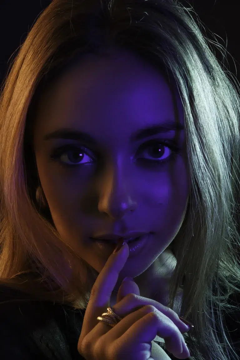 Close-up portrait of a woman with a finger on her lips, lit with violet light on the face and a warm light on her hair