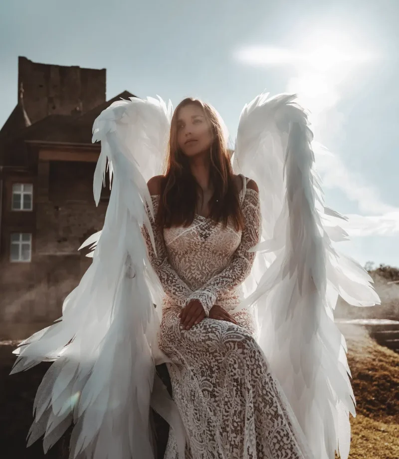 Outdoor fine art portrait of a woman in a lace gown with angel wings, photographed near a castle in soft sunlight.