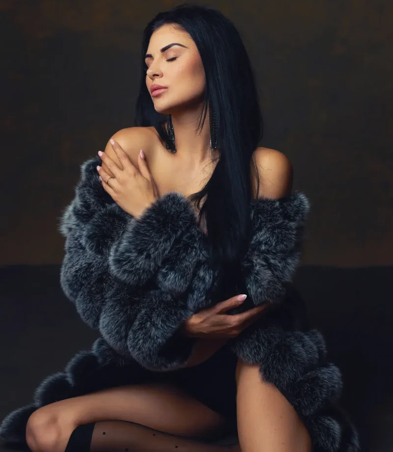 Boudoir portrait of a dark-haired woman wearing a grey fur coat, kneeling in a warm-toned studio, Massen Photography Luxembourg