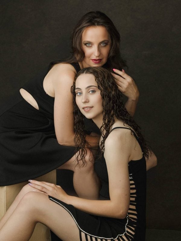 Studio portrait of mother and daughter wearing black dresses, sitting closely and smiling.