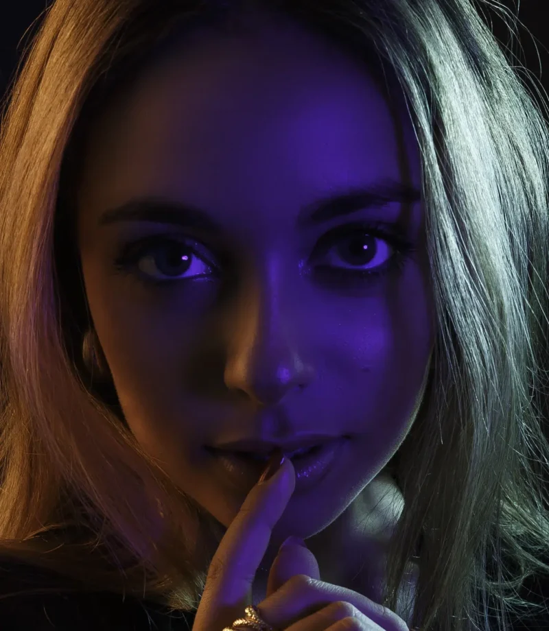 Close-up portrait of a woman with a finger on her lips, lit with violet light on the face and a warm light on her hair